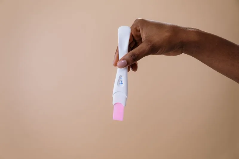 A hand holding a pregnancy test showing a plus sign in its window, against a beige background
