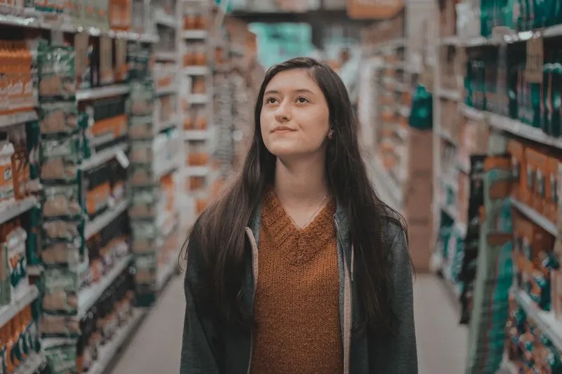 A thoughtful young woman standing in the middle of a shop aisle lined with shelves