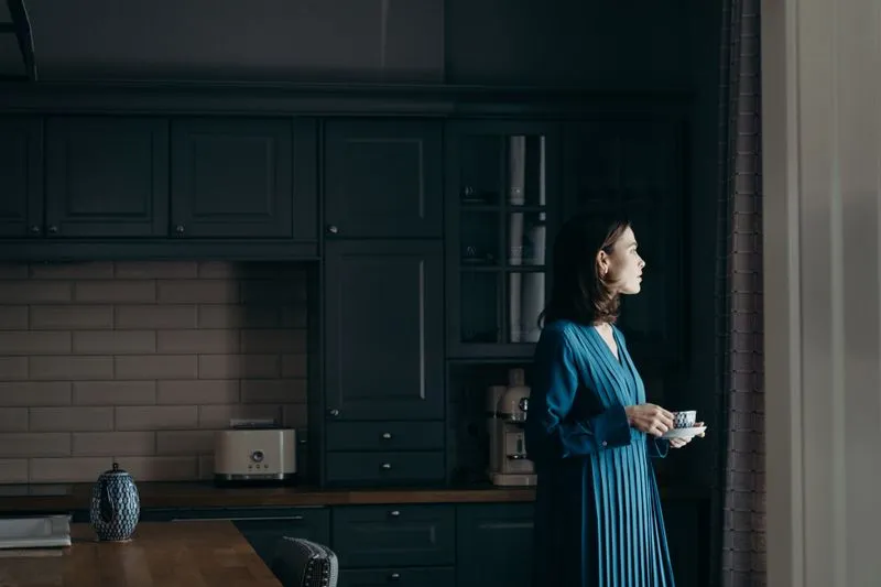 A woman standing in a dim kitchen, cup in hand, looking towards the window