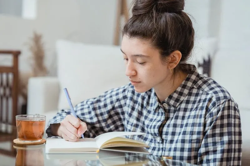 Young woman in a checked shirt writing in an open notebook, a glass of tea beside her