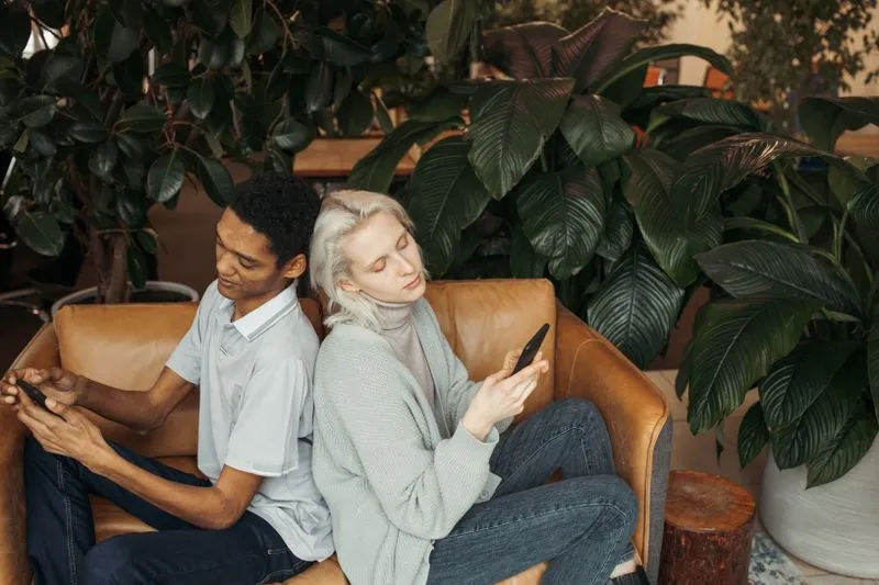 Young couple sitting back to back in a leather armchair, each absorbed in their own phone, surrounded by large green plants