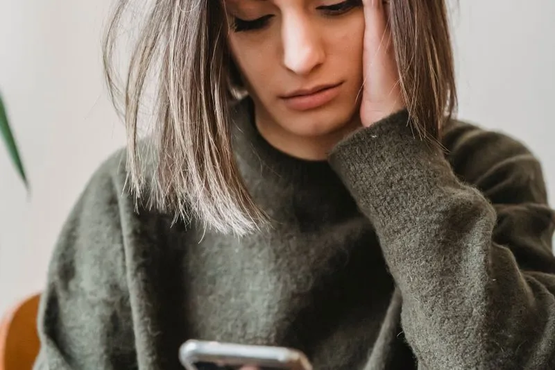 Young woman in an olive jumper, her head resting on her hand, eyes lowered to her phone, looking troubled