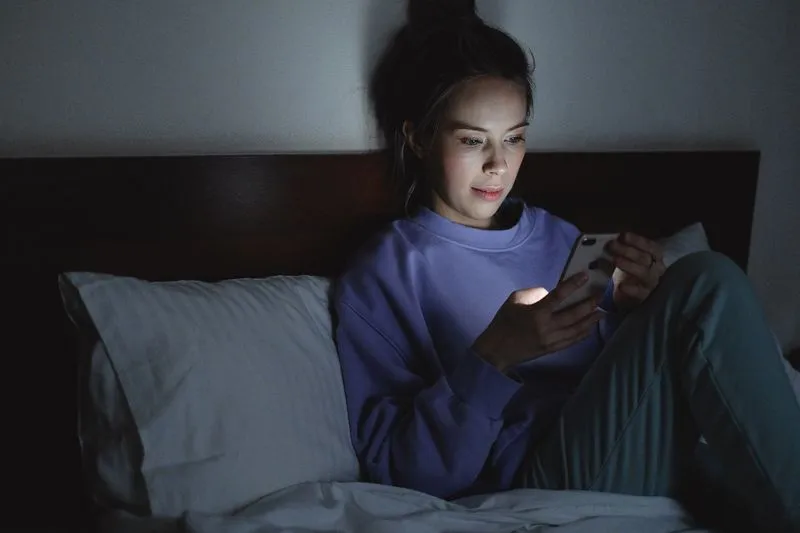 Young woman sitting up in bed at night, her face lit by the glow of her phone screen