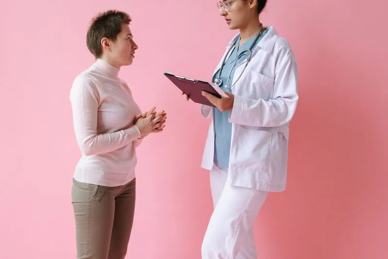 Young woman with short hair explaining her symptoms to a female doctor holding a clipboard