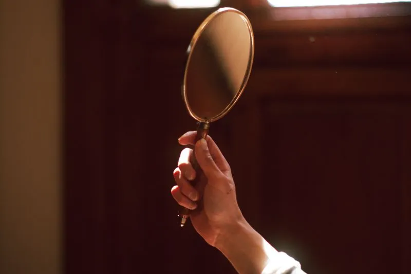 Hand holding an oval brass-rimmed hand mirror in a warmly lit room