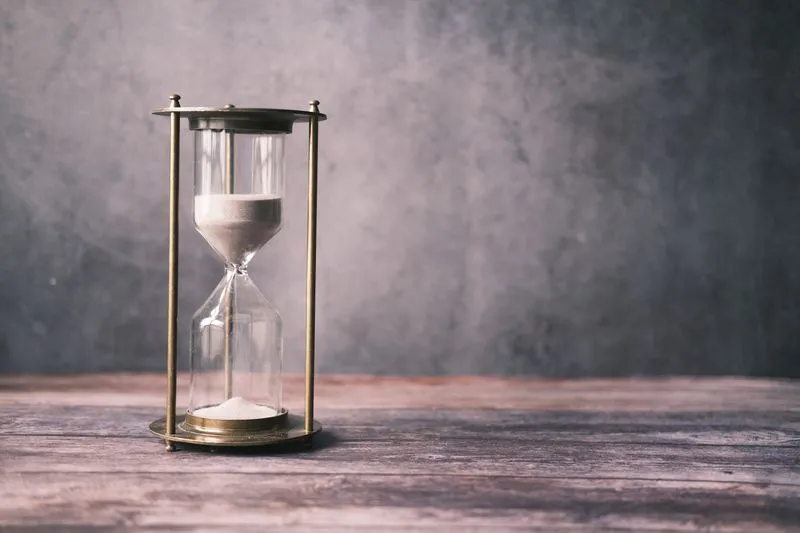 Brass-framed hourglass on a wooden table, white sand running into the lower bulb, against a grey wall