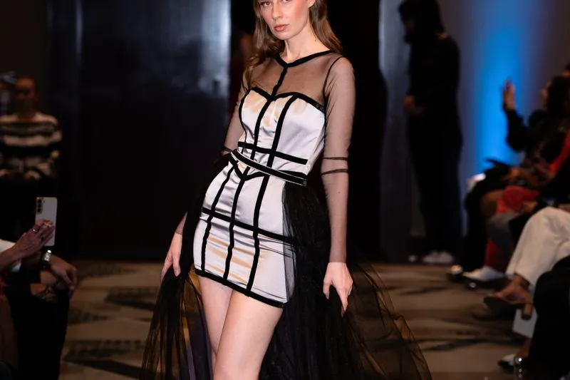 Model in a short grey satin dress with black bands and a tulle overskirt on a runway, with guests seated in the front row