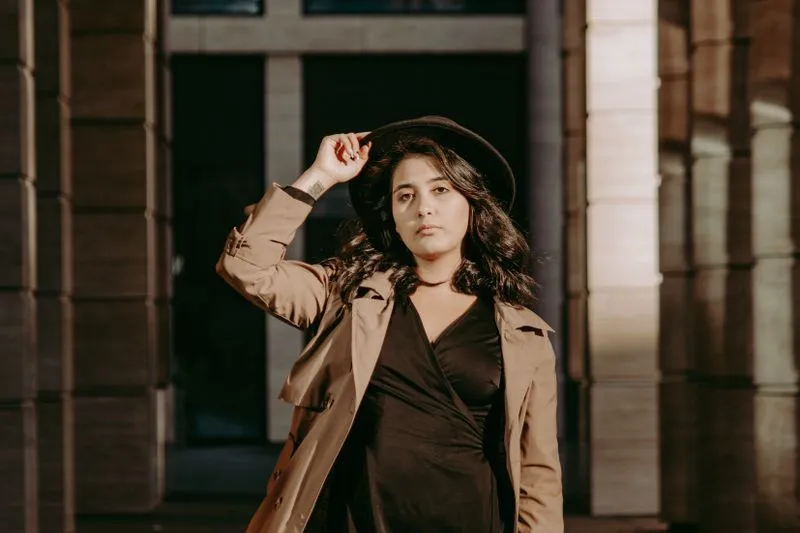 Woman in an open beige trench coat over a black wrap dress, wearing a black hat, between stone columns