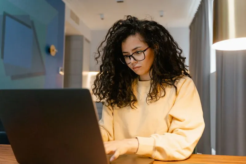 Woman with curly hair and glasses in a pale yellow sweatshirt, typing on a laptop