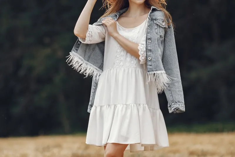 Woman in a short white ruffled dress with lace sleeves and a fringed denim jacket over her shoulders, in a field