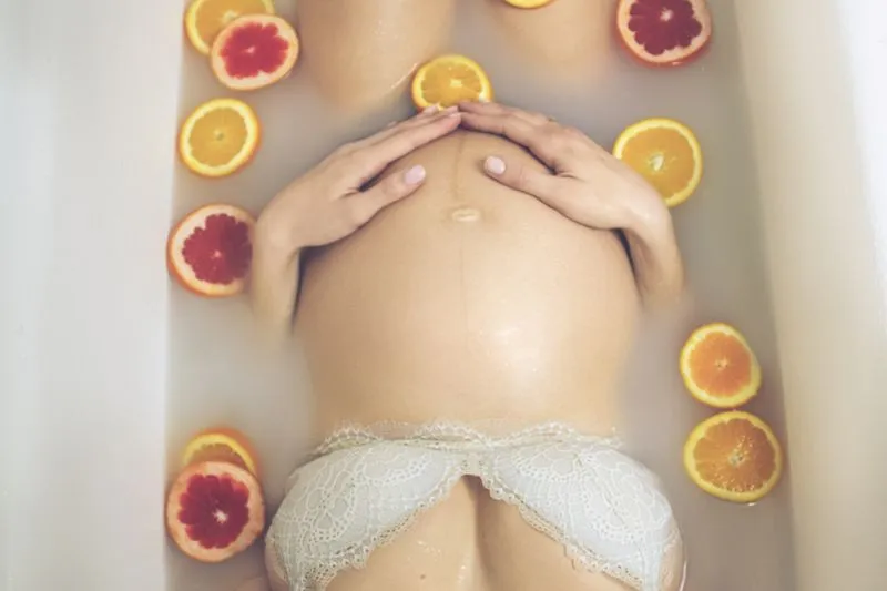 Pregnant woman lying in a milky bath with her hands on her bump, surrounded by orange and grapefruit slices
