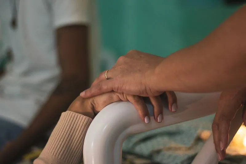 A hand resting on another person's hand as it grips the white rail of a hospital bed