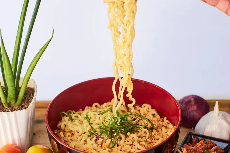 Instant noodles lifted with chopsticks above a red bowl, next to a small dish of kimchi