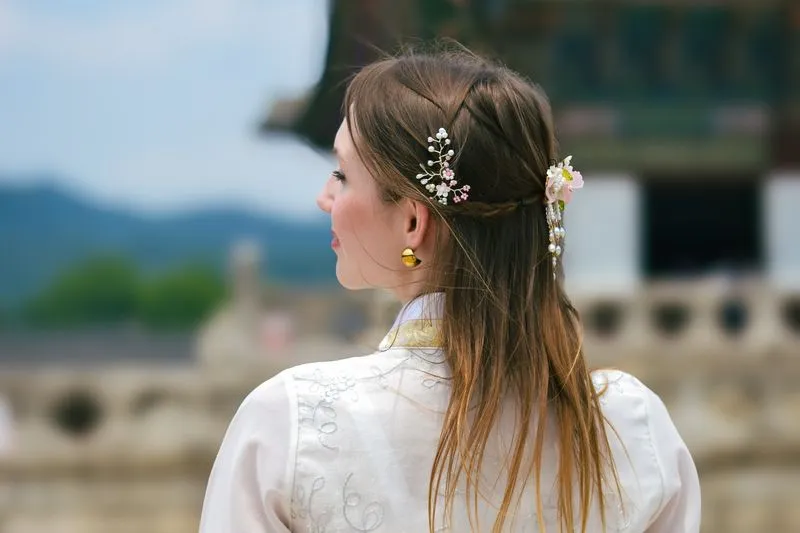 Young woman seen from behind in an embroidered white hanbok, flower ornaments in her hair, facing a Korean palace pavilion