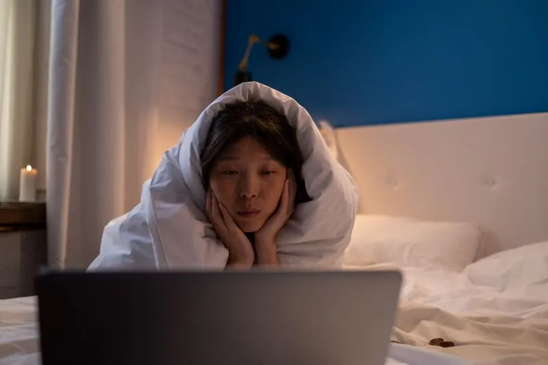 Young woman wrapped in a duvet on her bed, chin in her hands, watching a laptop screen in the evening