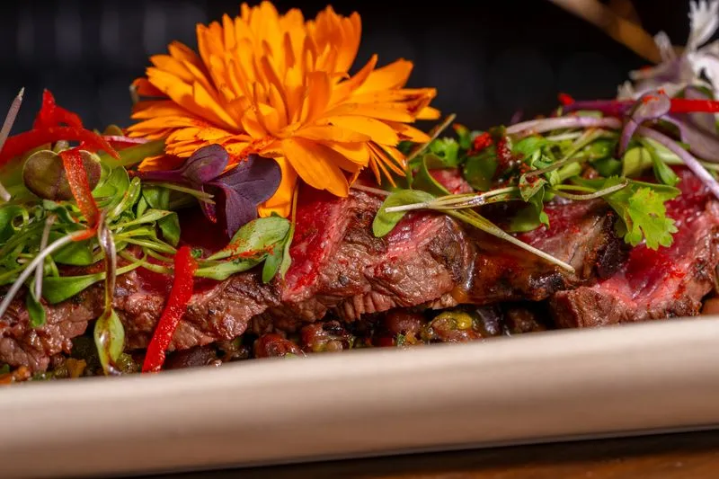 Slices of rare beef plated with an orange edible flower, microgreens and strips of red pepper
