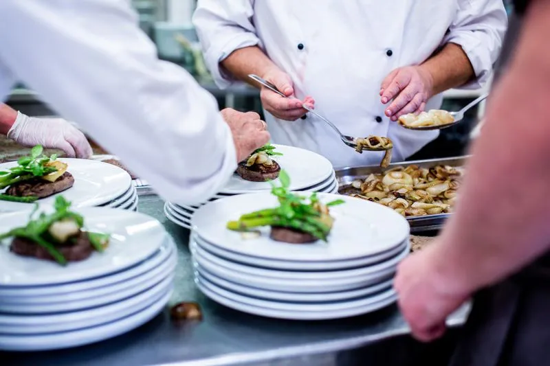 Cooks in white jackets plating rows of identical dishes topped with green asparagus, on stacks of plates