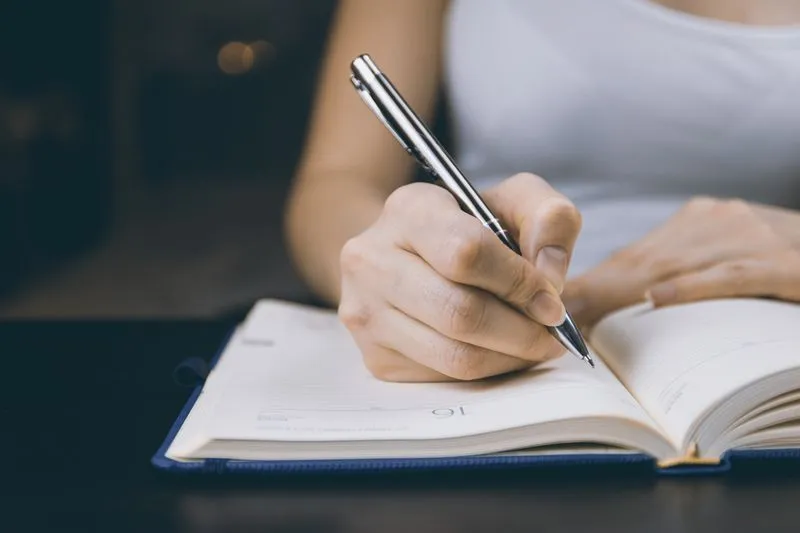 Hand writing with a silver pen in an open diary with a navy cover, on a dark table