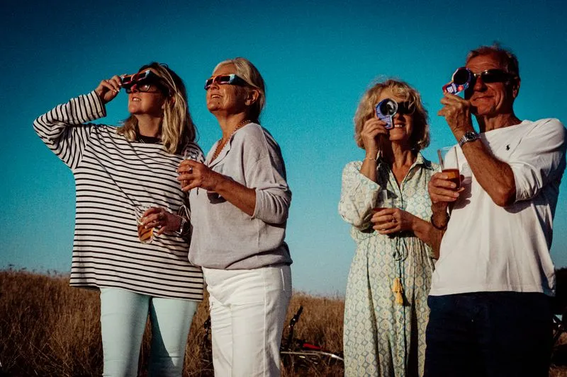 Four adults outdoors, faces turned to the sky, watching an eclipse through cardboard glasses and hand-held viewers