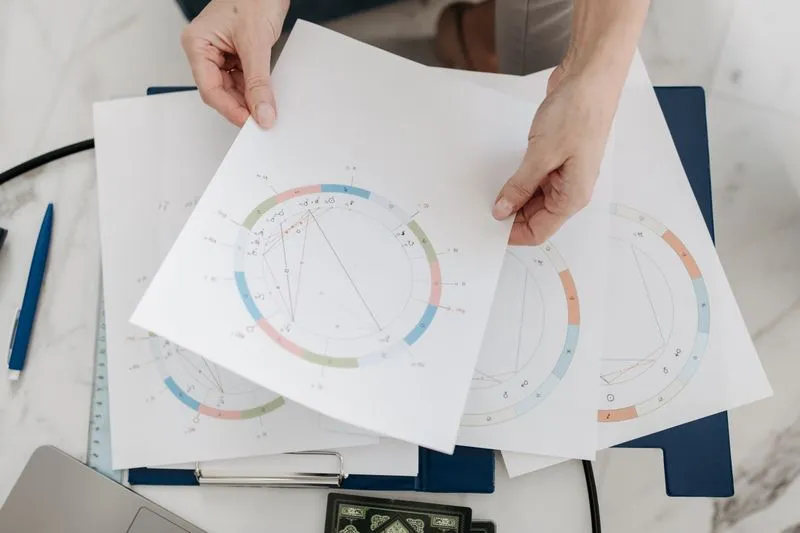 Hands holding several printed birth charts with colour-coded house wheels above a desk