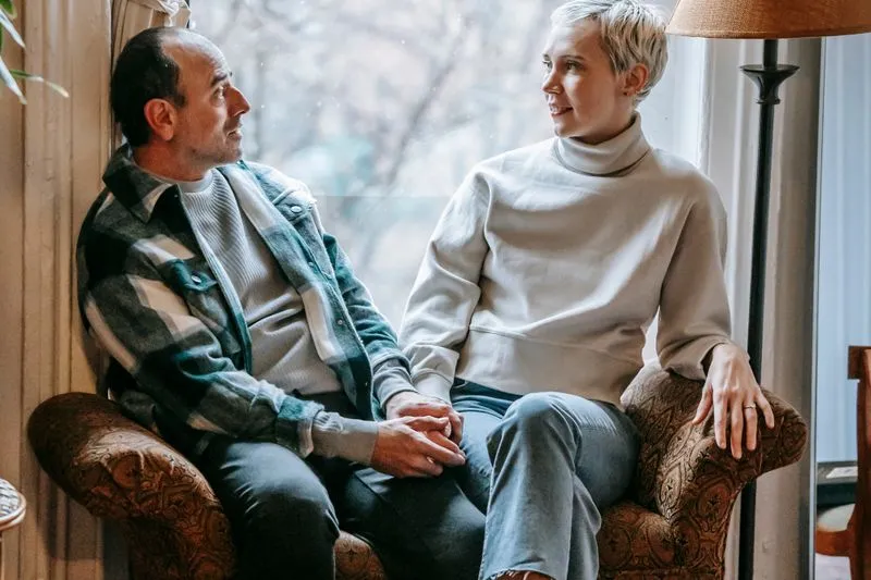 A couple sitting in an armchair by a window, holding hands and talking calmly