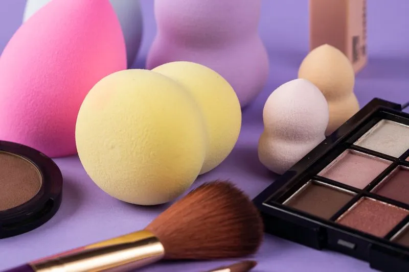 Make-up sponges in assorted shapes and colours on a purple background, with a brush, an eyeshadow palette and a pressed powder