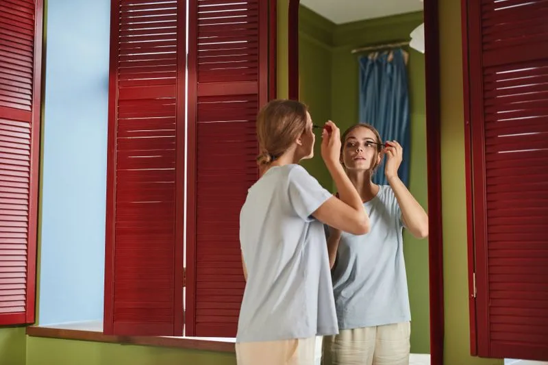 Young woman running a mascara wand through her lashes in front of a mirror