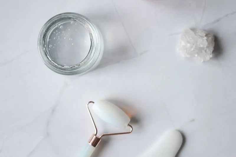 Open glass jar of clear gel on marble, next to a facial roller