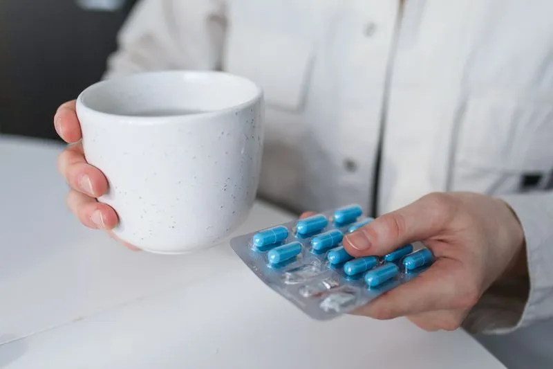 Hands holding a white mug and a blister pack of blue capsules above a table