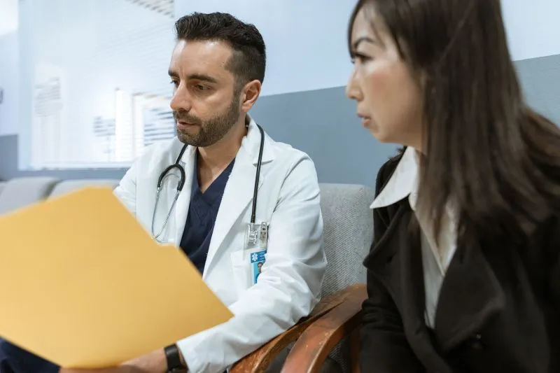 A doctor in a white coat, stethoscope round his neck, going through a yellow folder beside an attentive patient