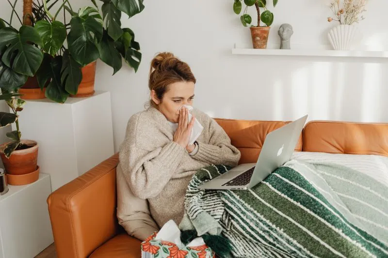 A woman on a sofa under a throw, blowing her nose in front of her laptop, with a box of tissues at her feet