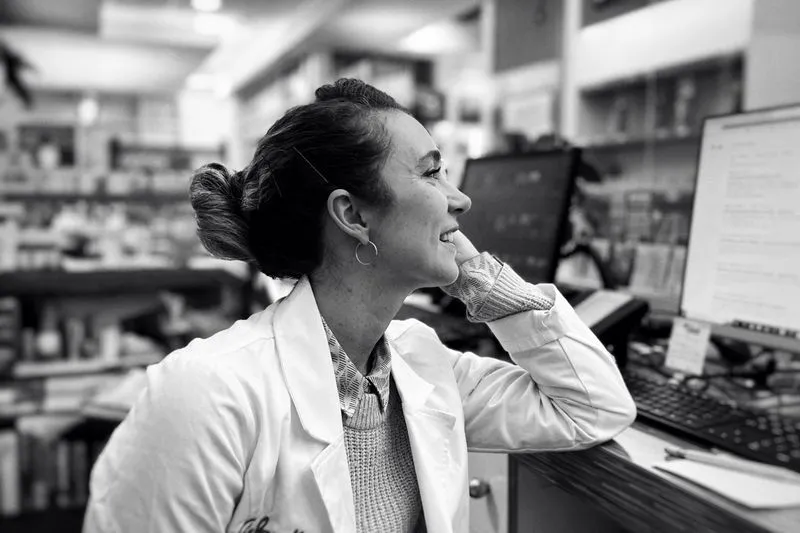 A smiling pharmacist in a white coat leaning on her counter next to her computer screens, in black and white