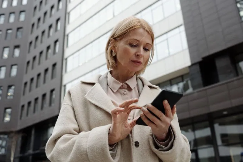A woman in a pale coat checking her smartphone on a street lined with office buildings