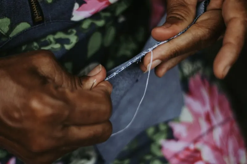 Hands sewing the edge of a grey fabric with a needle, a white thread pulled taut
