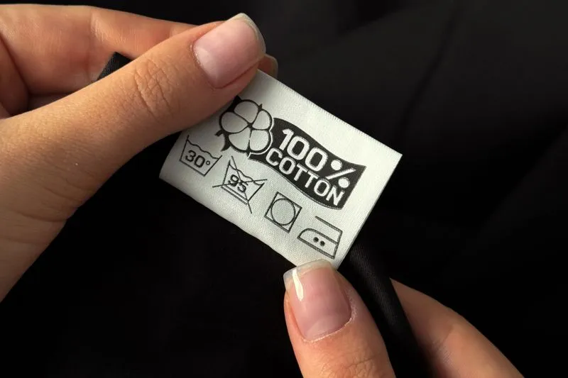 Hands holding a sewn-in 100% cotton label with care symbols on a black garment