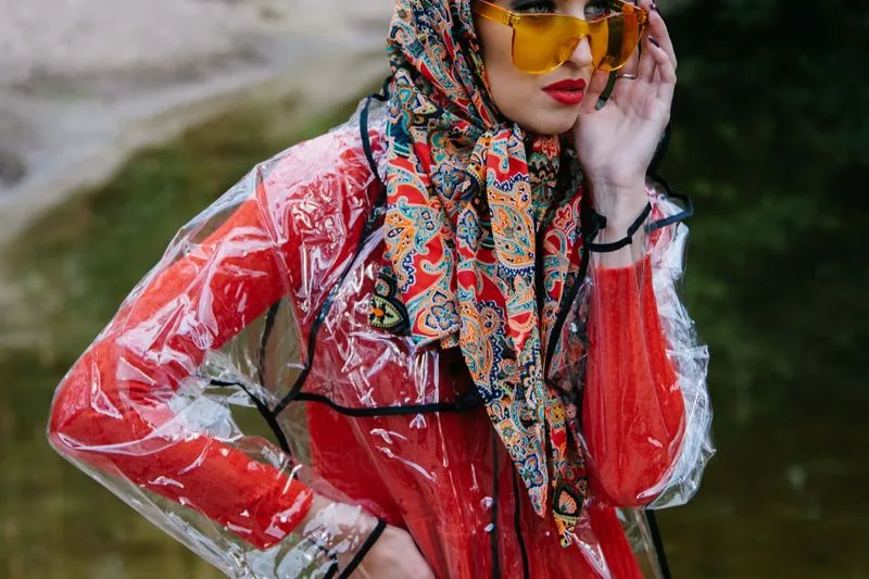 Woman with a paisley-print scarf over her head, yellow sunglasses and a clear raincoat over a red dress