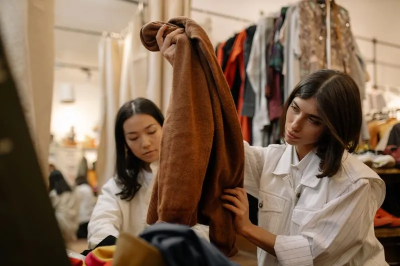 Two women examining a brown sweater in a second-hand shop, in front of clothing racks