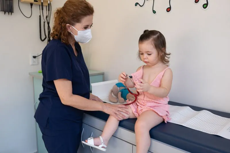 Little girl of about two sitting on an examination table, playing with a stethoscope next to a masked doctor who rests a hand on her knee