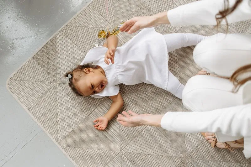 Little girl lying on her back on a rug, crying and screaming, while an adult crouches and reaches out to her
