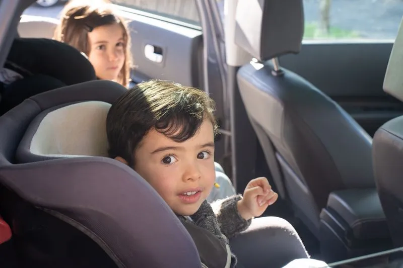 Little boy sitting in his car seat, the car parked with the door open, a girl further back
