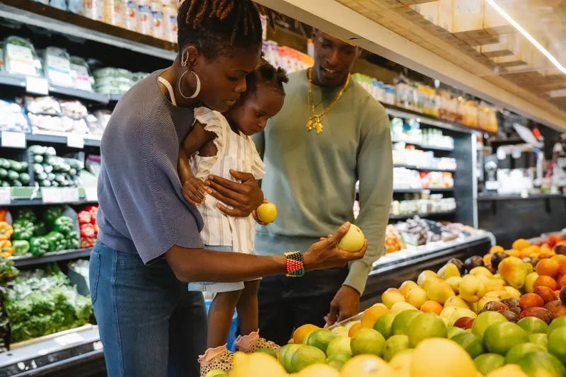 Mother carrying her little girl on her hip by the fruit display in a supermarket, the child holding an apple, the father beside them