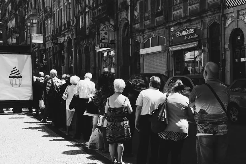 Black-and-white photo of a long queue of people waiting on a sunny pavement in front of old shopfronts