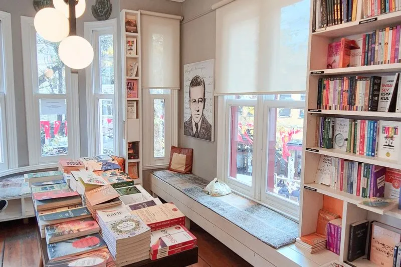Corner of a bright bookshop: a table stacked with books, full white shelves and a window seat under tall windows