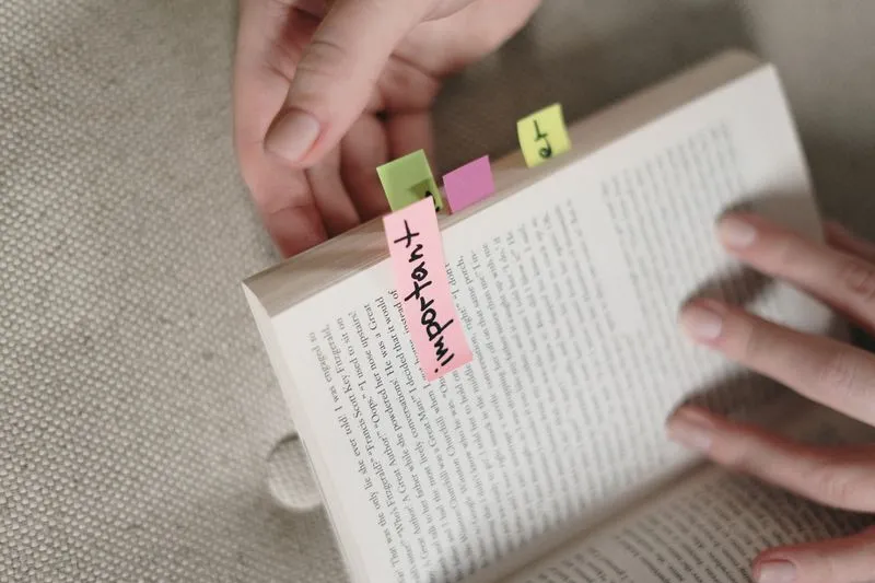 Open novel held in both hands, marked with small coloured sticky notes, a pink one reading ‘important’