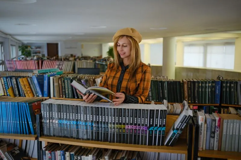 Reader in a beret and checked jacket leafing through an open book between shelves full of books