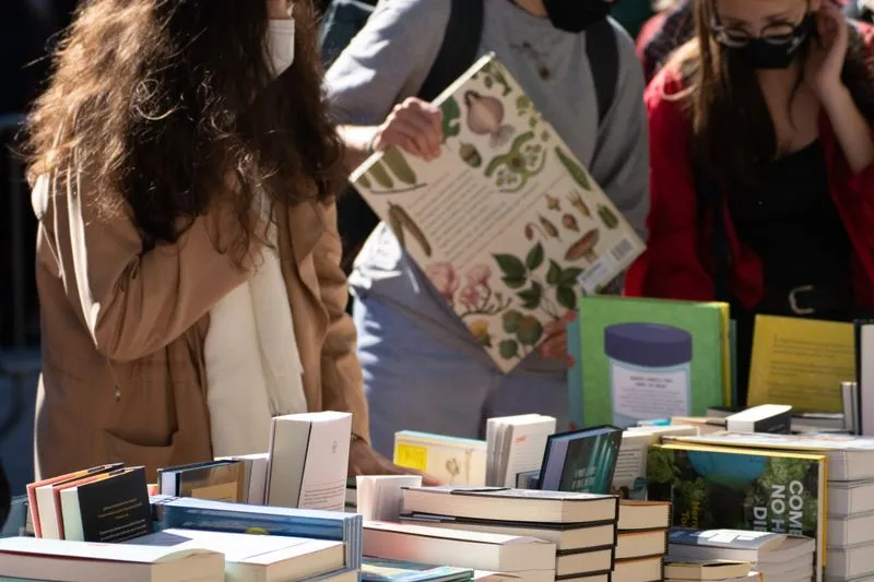 Visitors browsing books around a table at an outdoor book fair, novels and illustrated books stacked up