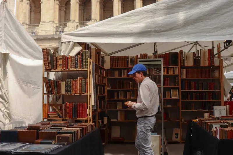 Stall under a white tent at an outdoor book fair, shelves of bound books and a visitor in a cap leafing through one