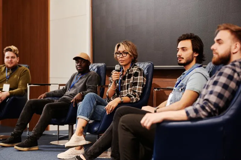 Five speakers seated in armchairs on a stage, a woman in glasses speaking into a microphone