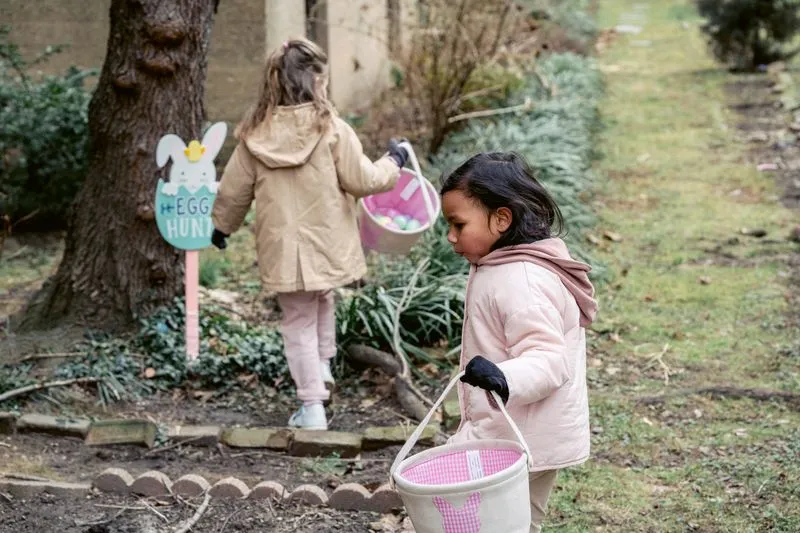 Two little girls in coats, Easter baskets in hand, looking for eggs in a garden beside an “Egg hunt” sign