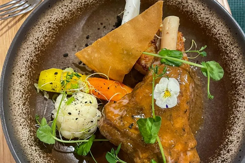 Braised lamb shank coated in its juices, served with carrots, cauliflower and pea shoots